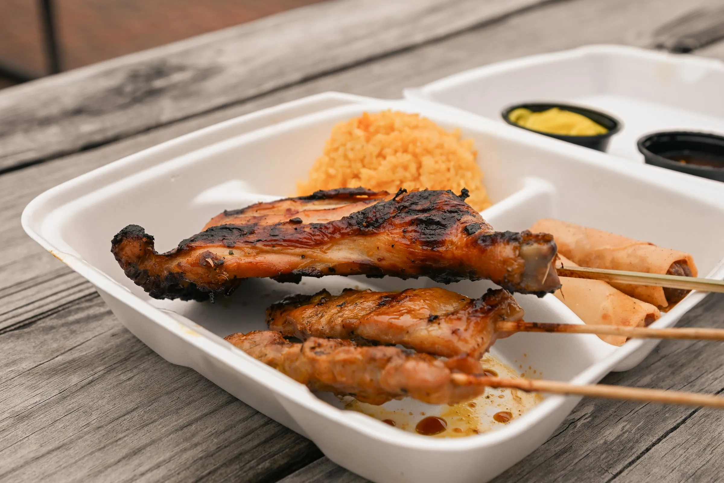 Close view of a combo plate with a grilled chicken leg, pork barbecue skewers, lumpia, and java rice
