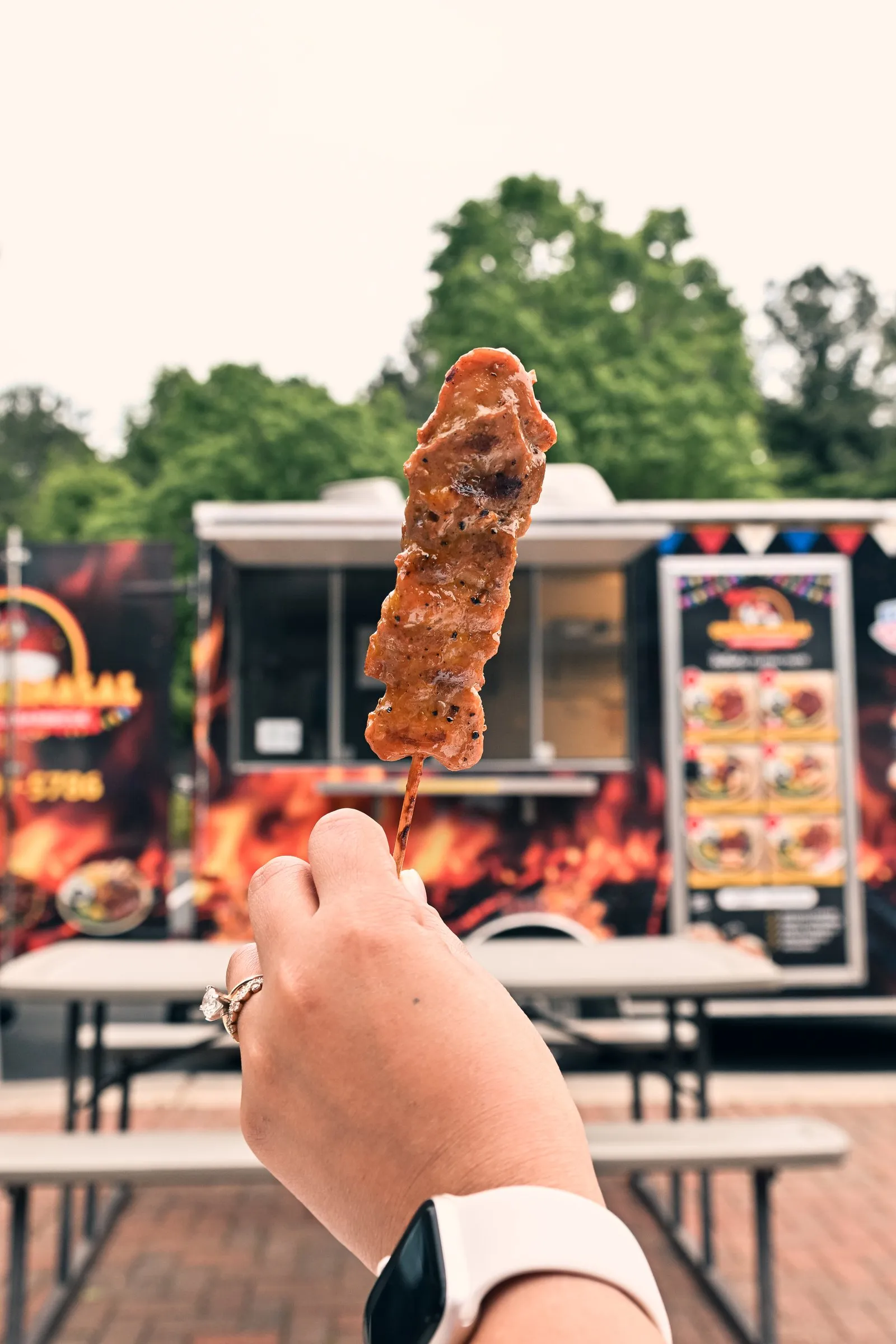A barbecue skewer held up over the picnic tables with the food truck and menu board behind it