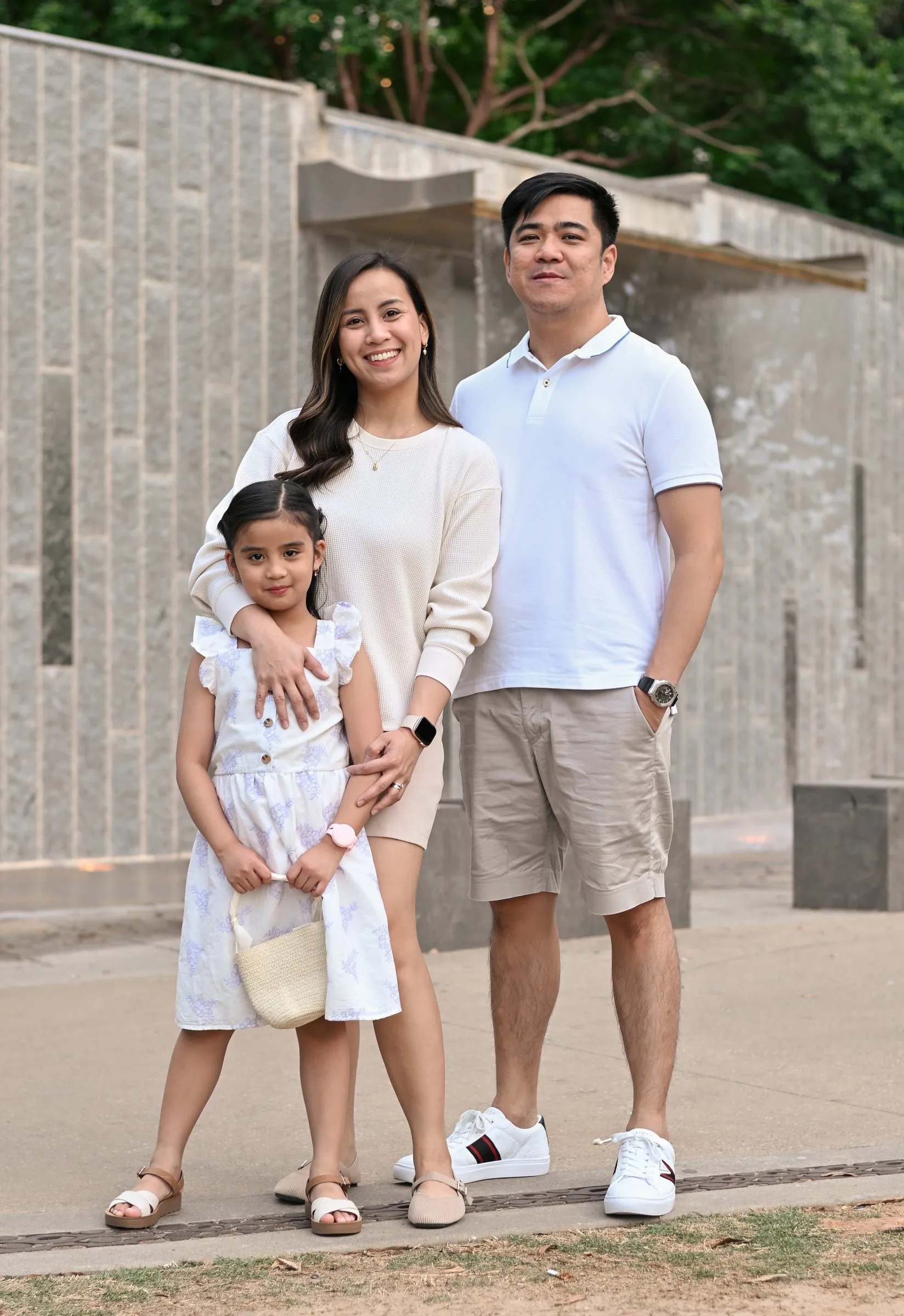 Jessica, Jet, and their daughter in cream and white standing in front of the stone wall at Romare Bearden Park