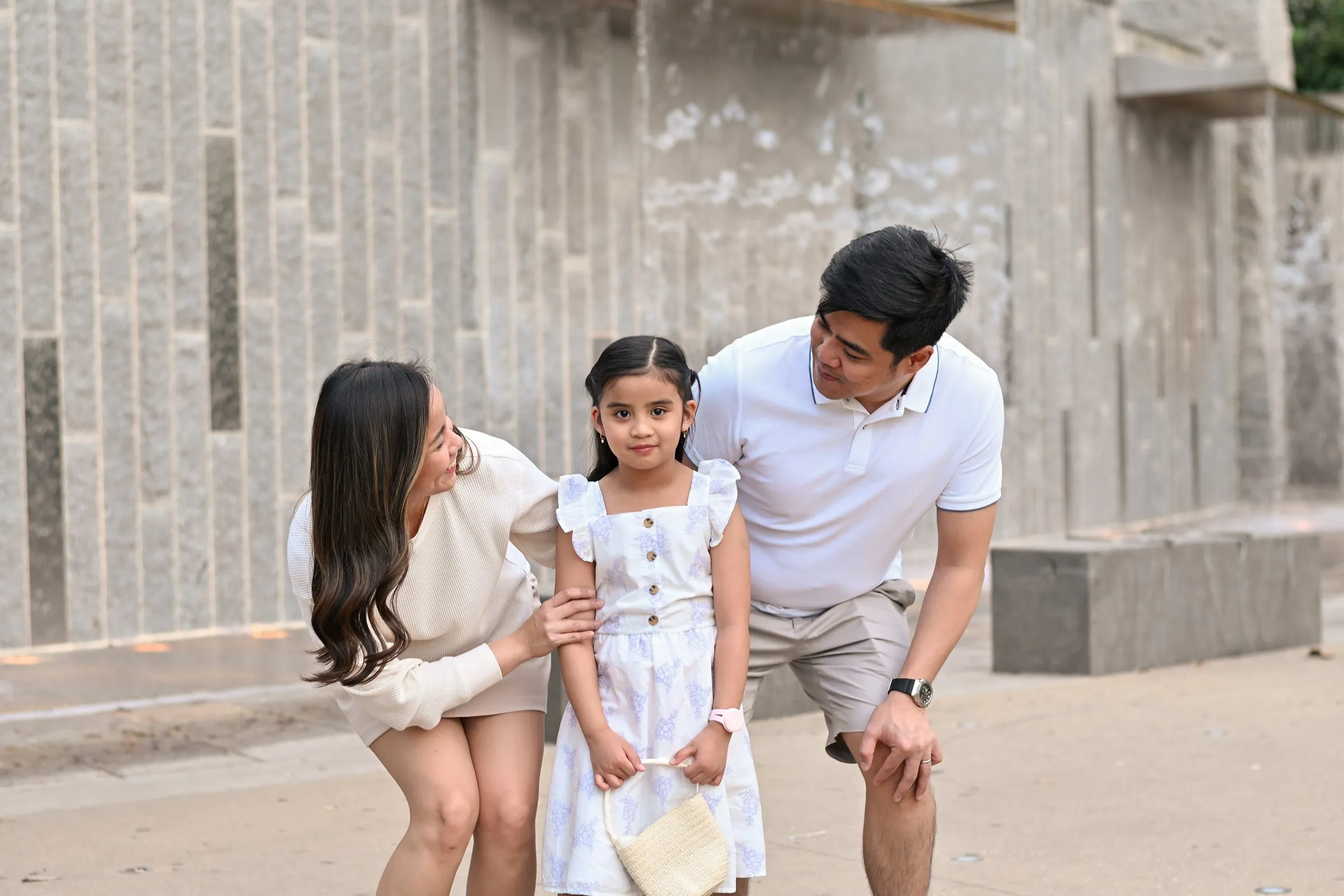 Jessica and Jet lean in toward their daughter, all three laughing, in front of the stone wall