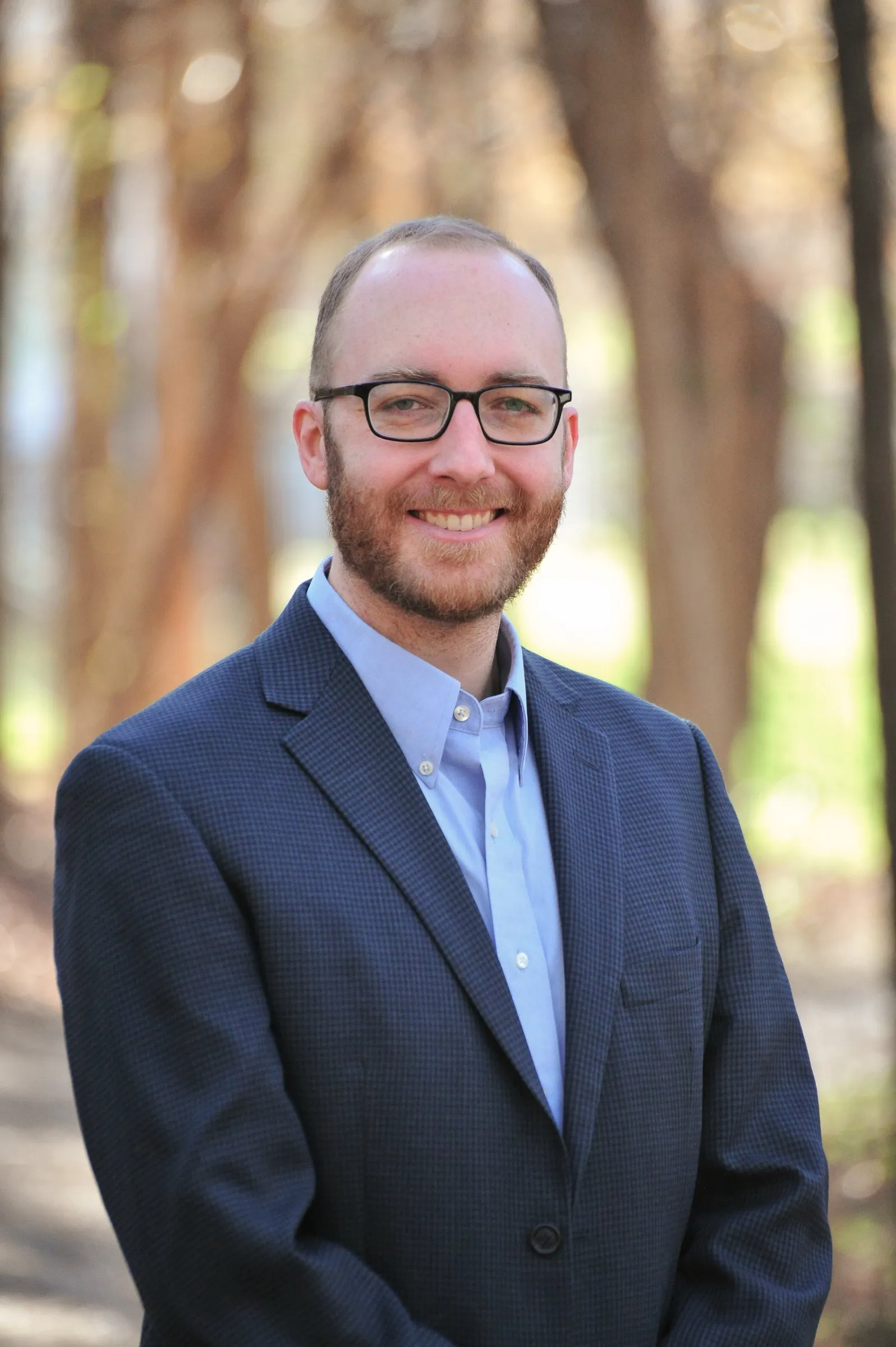 Portrait of Matt in a navy blazer and light blue shirt, smiling under the trees