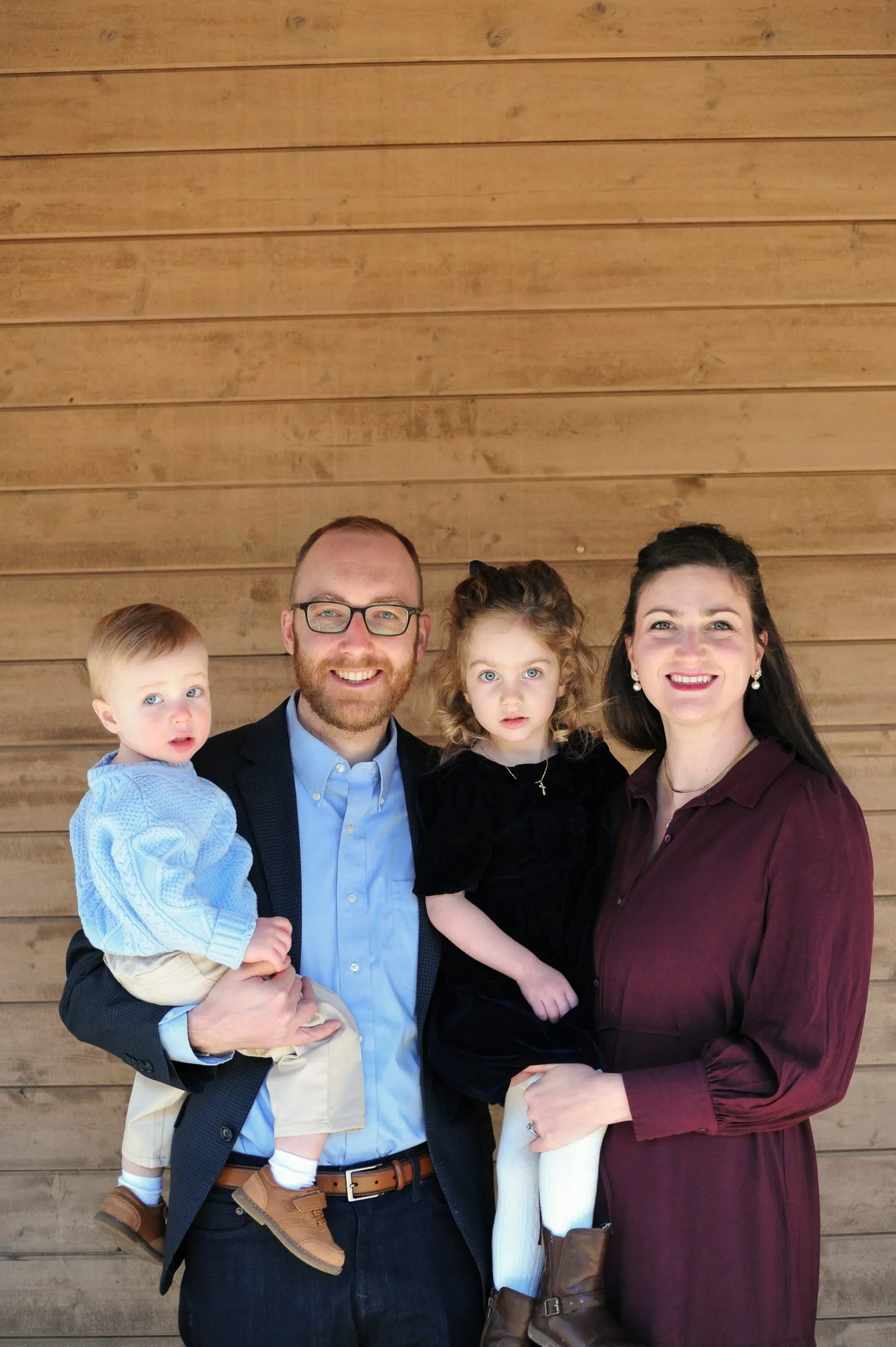 Matt holding their son and Olivia holding their daughter in front of a wood-sided wall