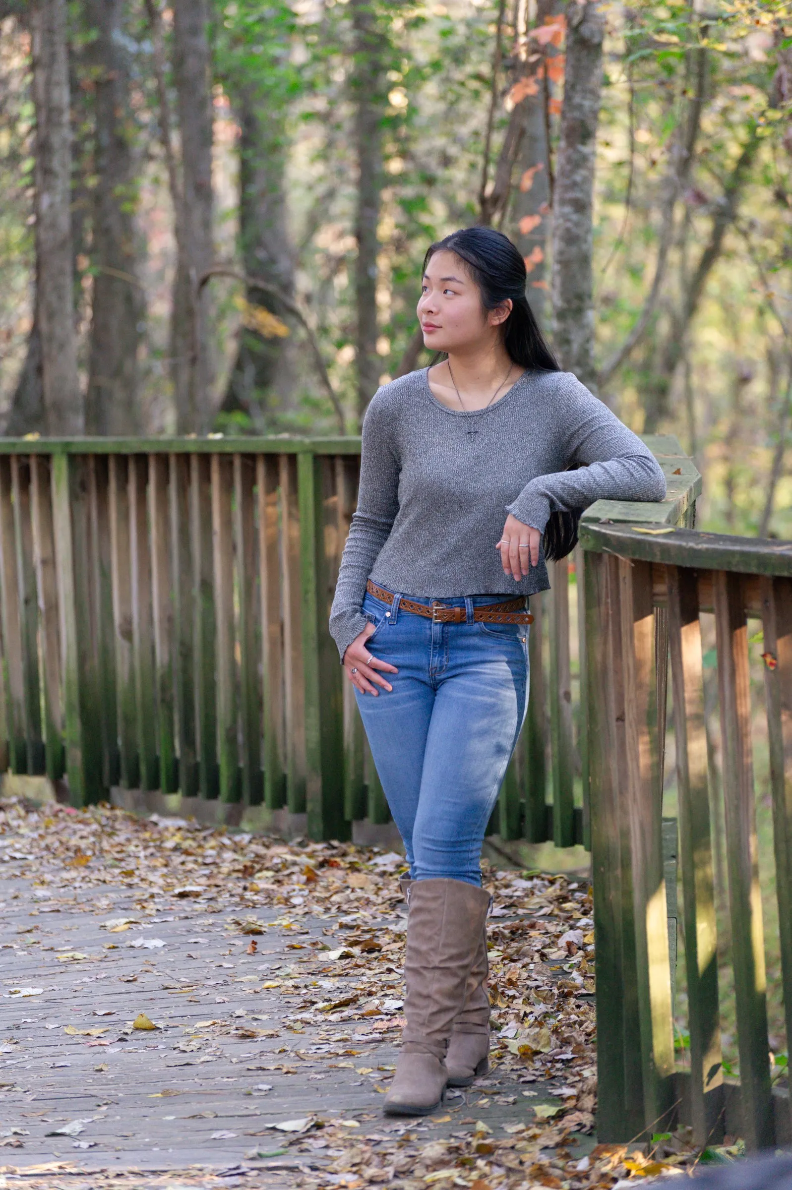Meilin leaning on a wooden boardwalk railing in a gray sweater and tall boots, fallen leaves at her feet