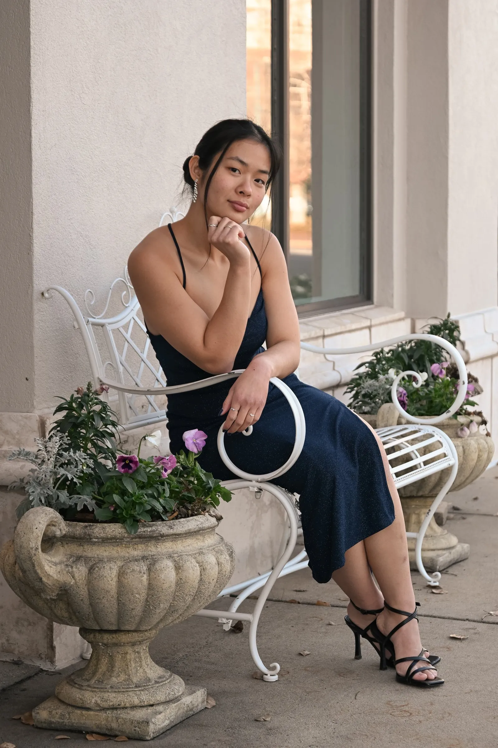 Meilin resting her chin on her hand on the white iron bench, strappy black heels and pansies beside her