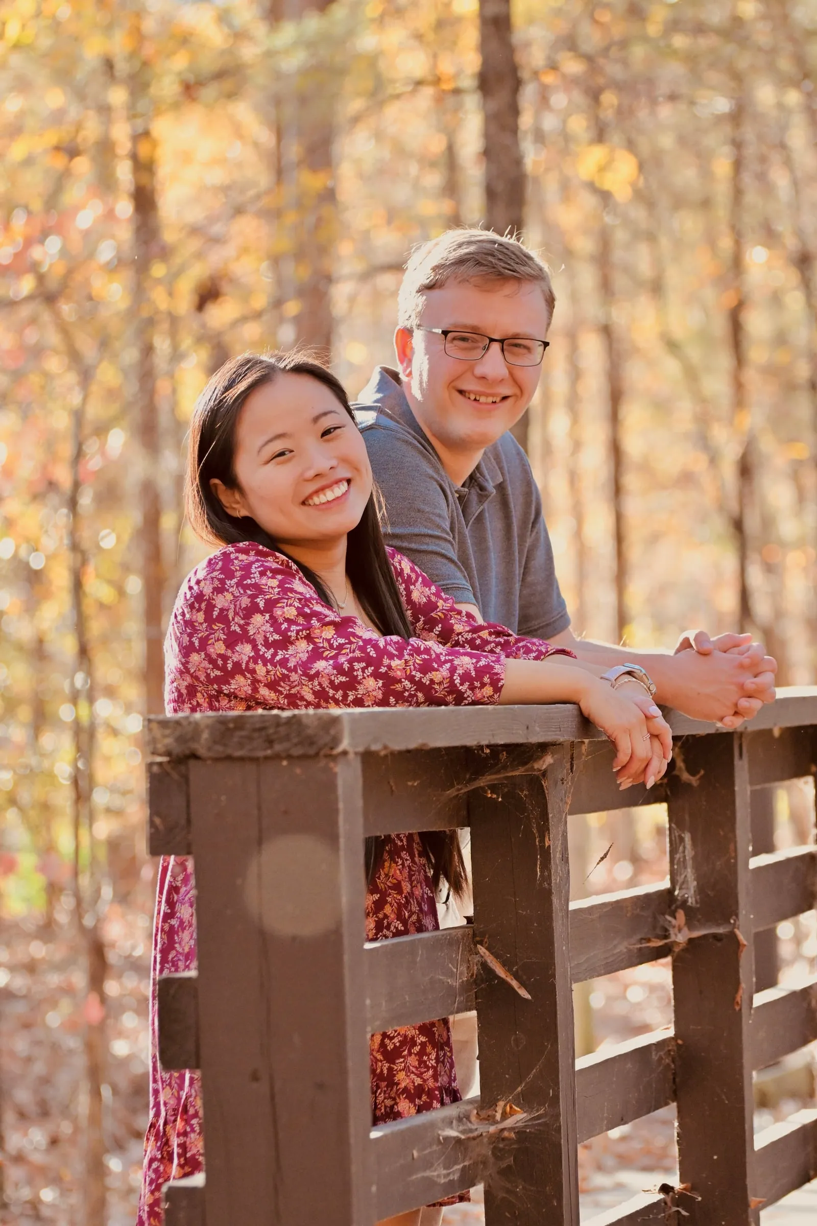 Mya and Jacob leaning on a wooden rail and smiling at the camera, golden fall trees behind them