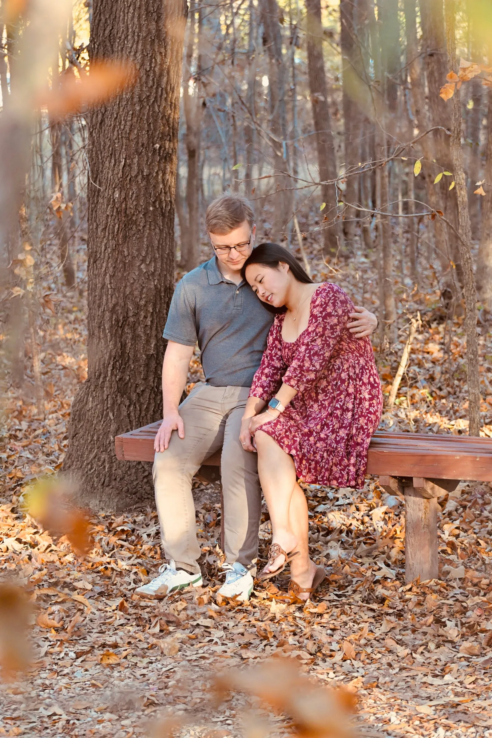 Mya resting her head on Jacob's shoulder on a wooden bench under a tall pine, fallen leaves in the foreground