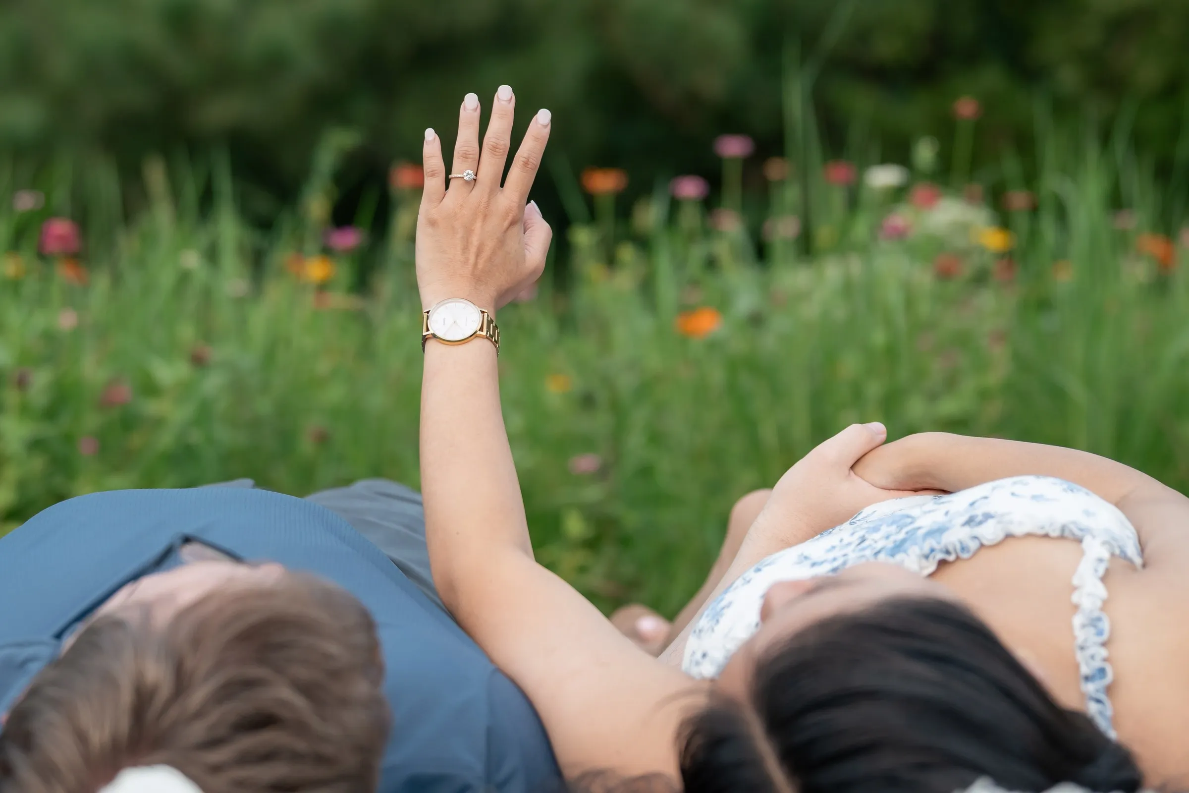 Mya raising her hand to show her engagement ring as the couple lies in the grass