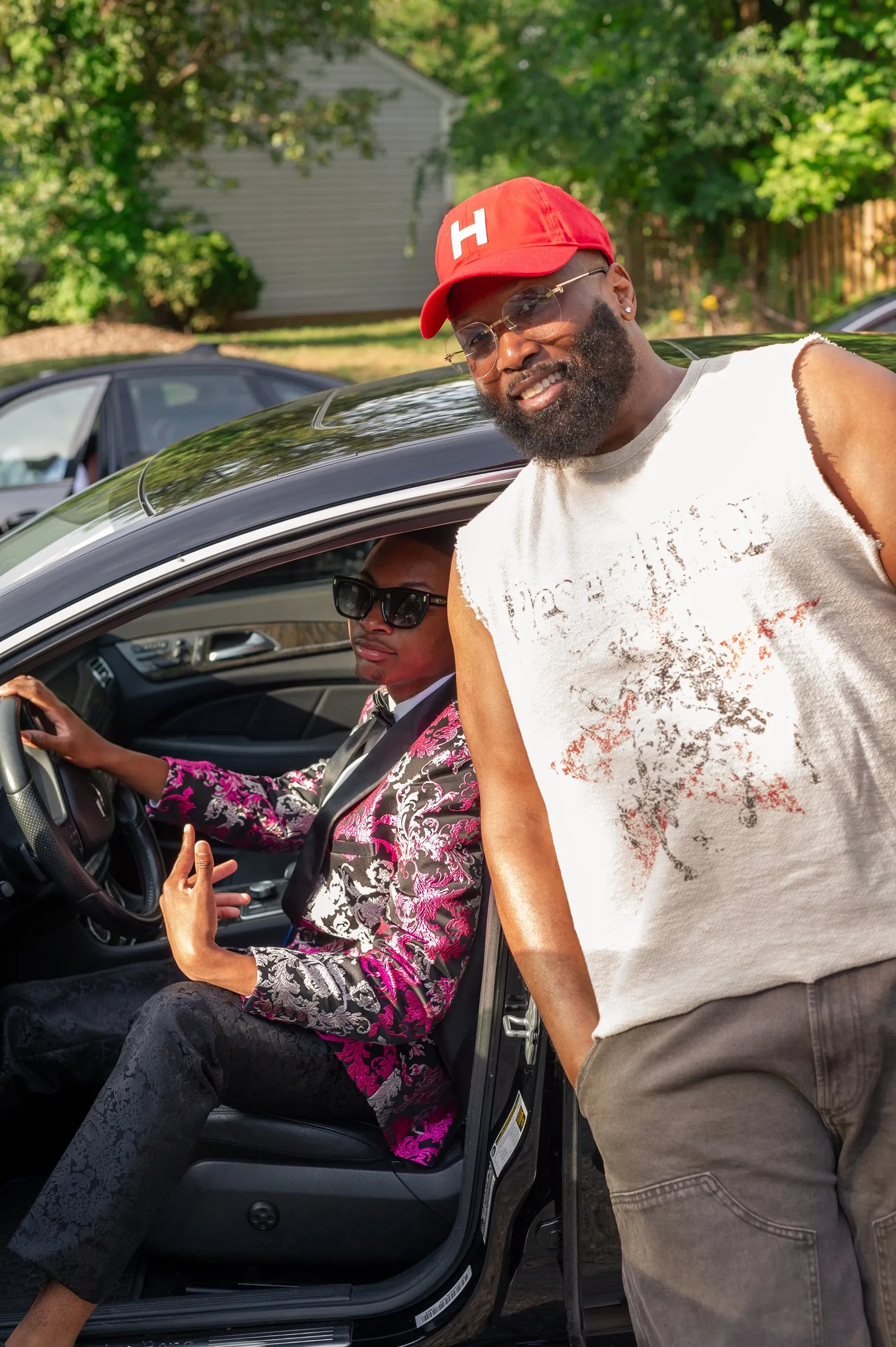He sits in the driver's seat of a black Mercedes as a man in a red cap leans in beside him