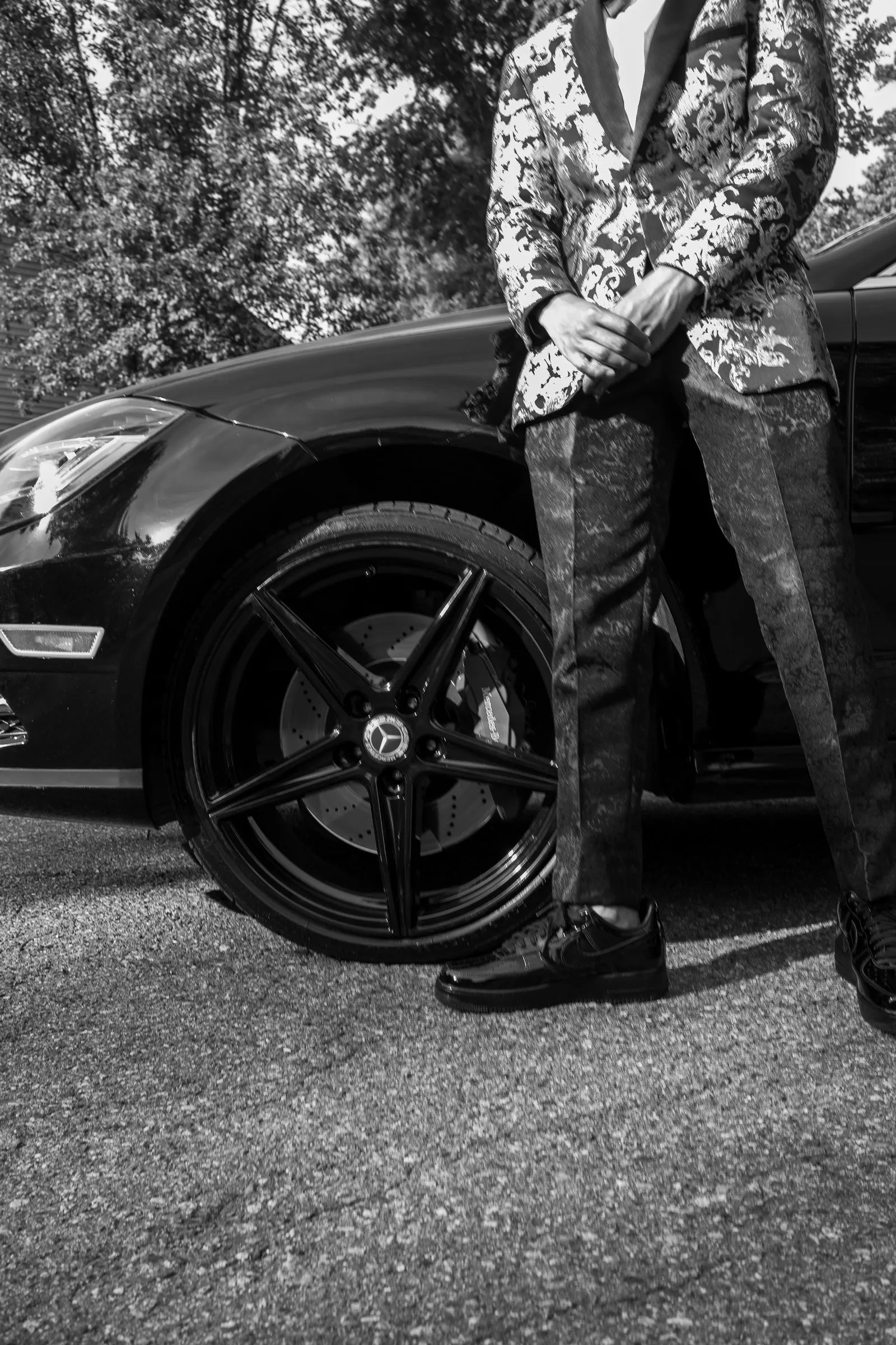 Black and white detail of his patterned trousers and dress shoes beside the Mercedes wheel