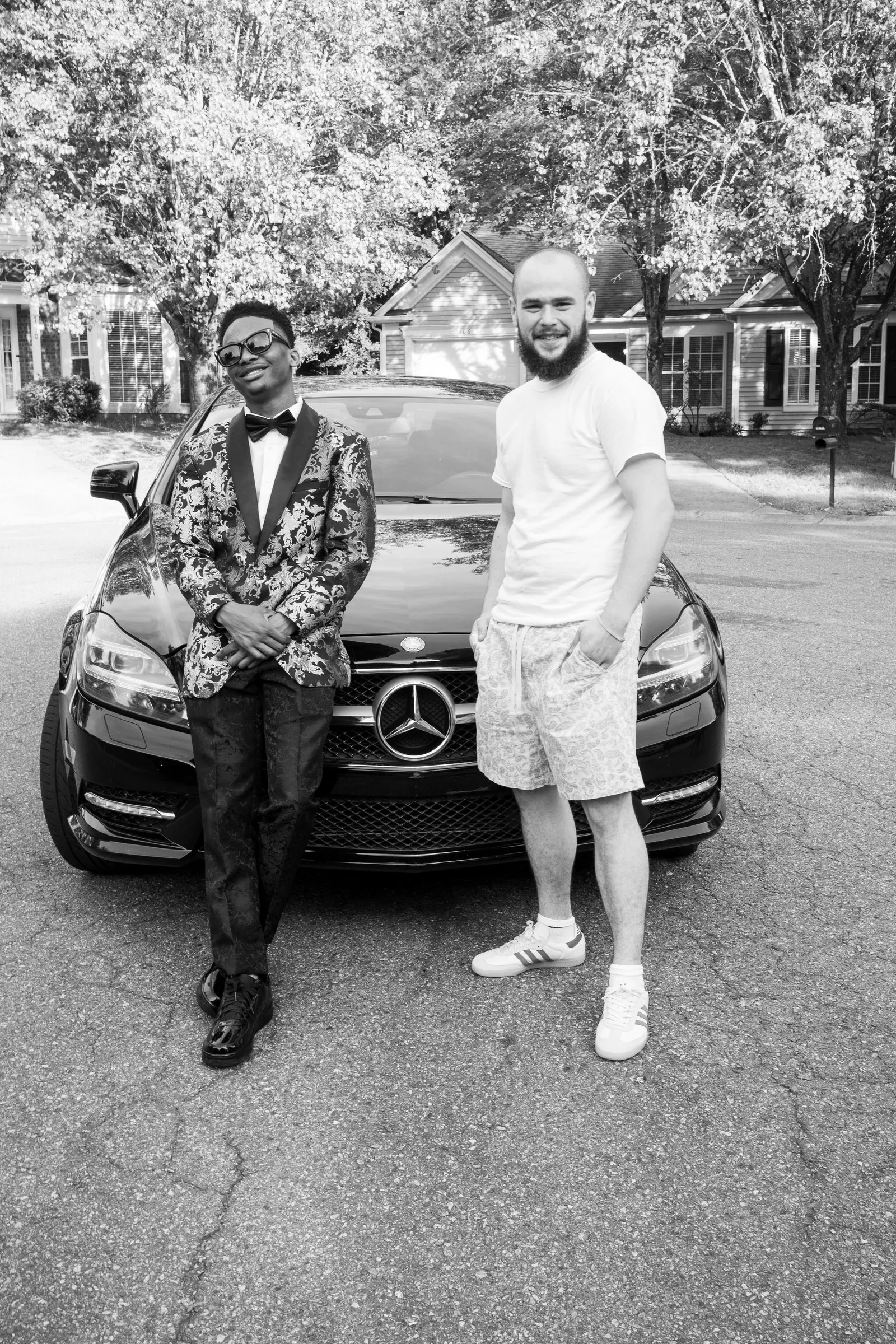 Black and white photograph of him with a man in a white tee in front of the Mercedes
