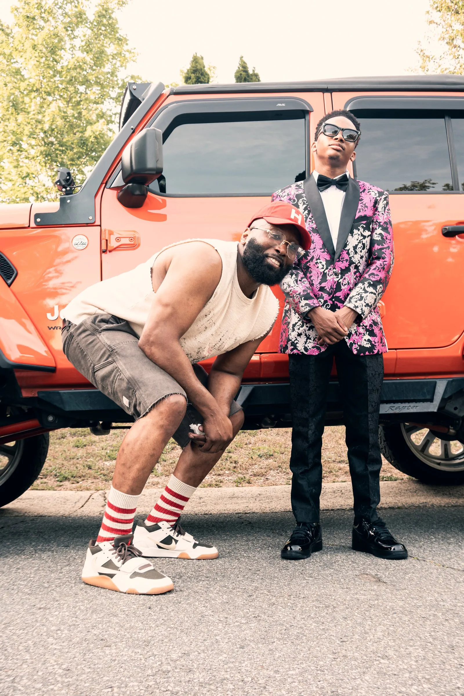 A man in a red cap crouches beside him in front of the orange Jeep