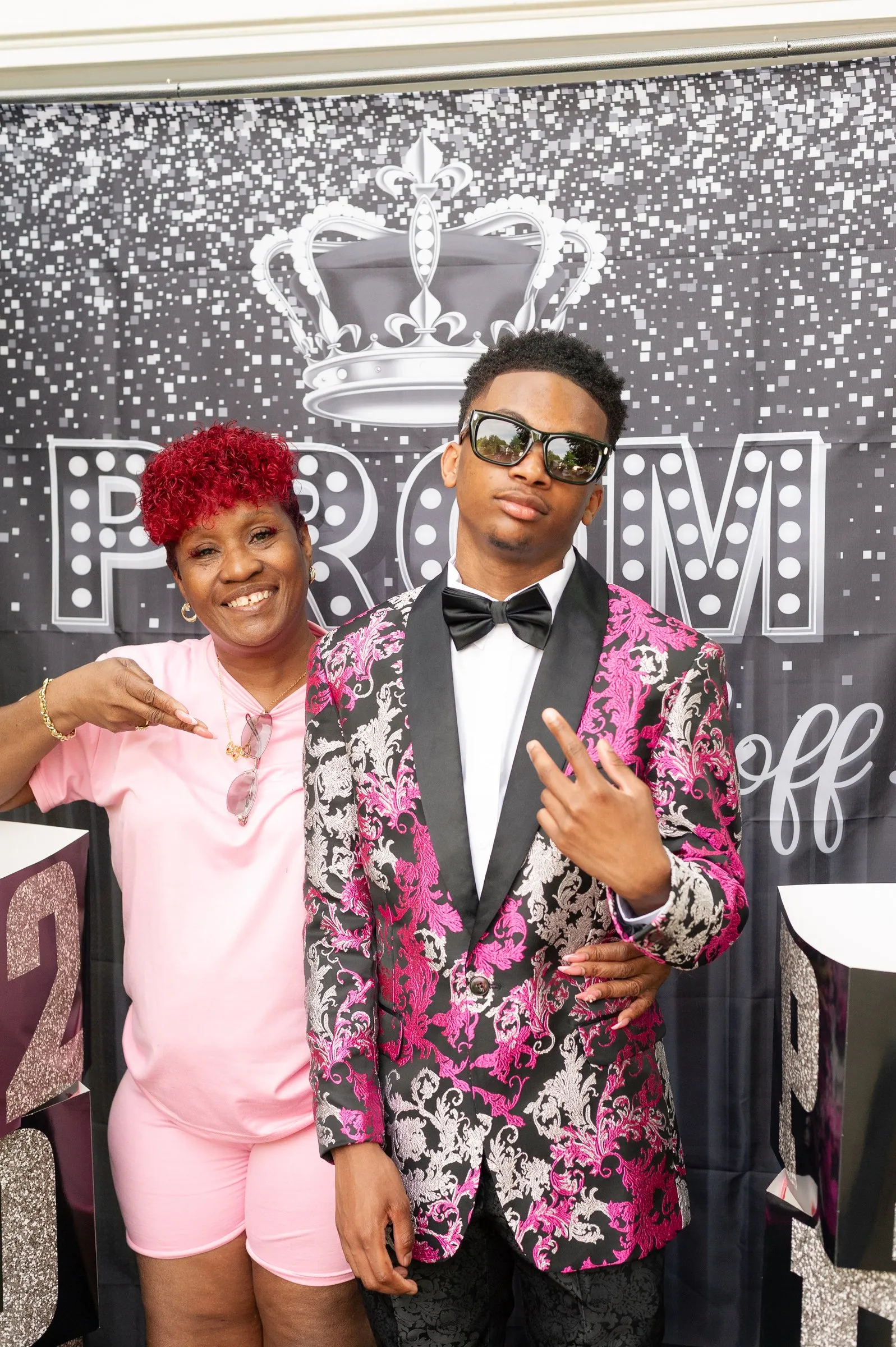 He poses with a woman in pink in front of the glitter Prom Send Off backdrop with a silver crown