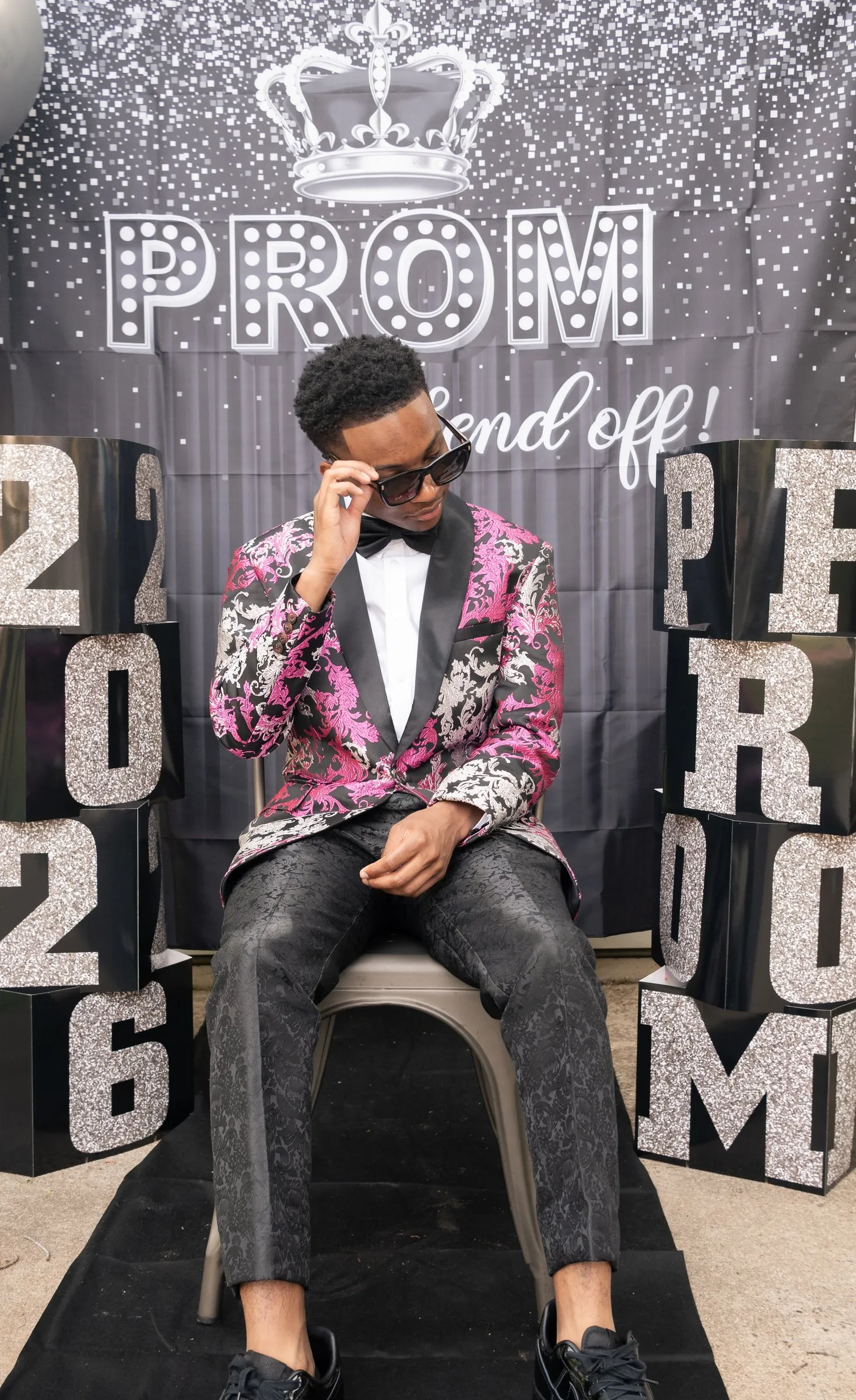 He sits on the black runner lowering his sunglasses, between the 2026 and PROM blocks