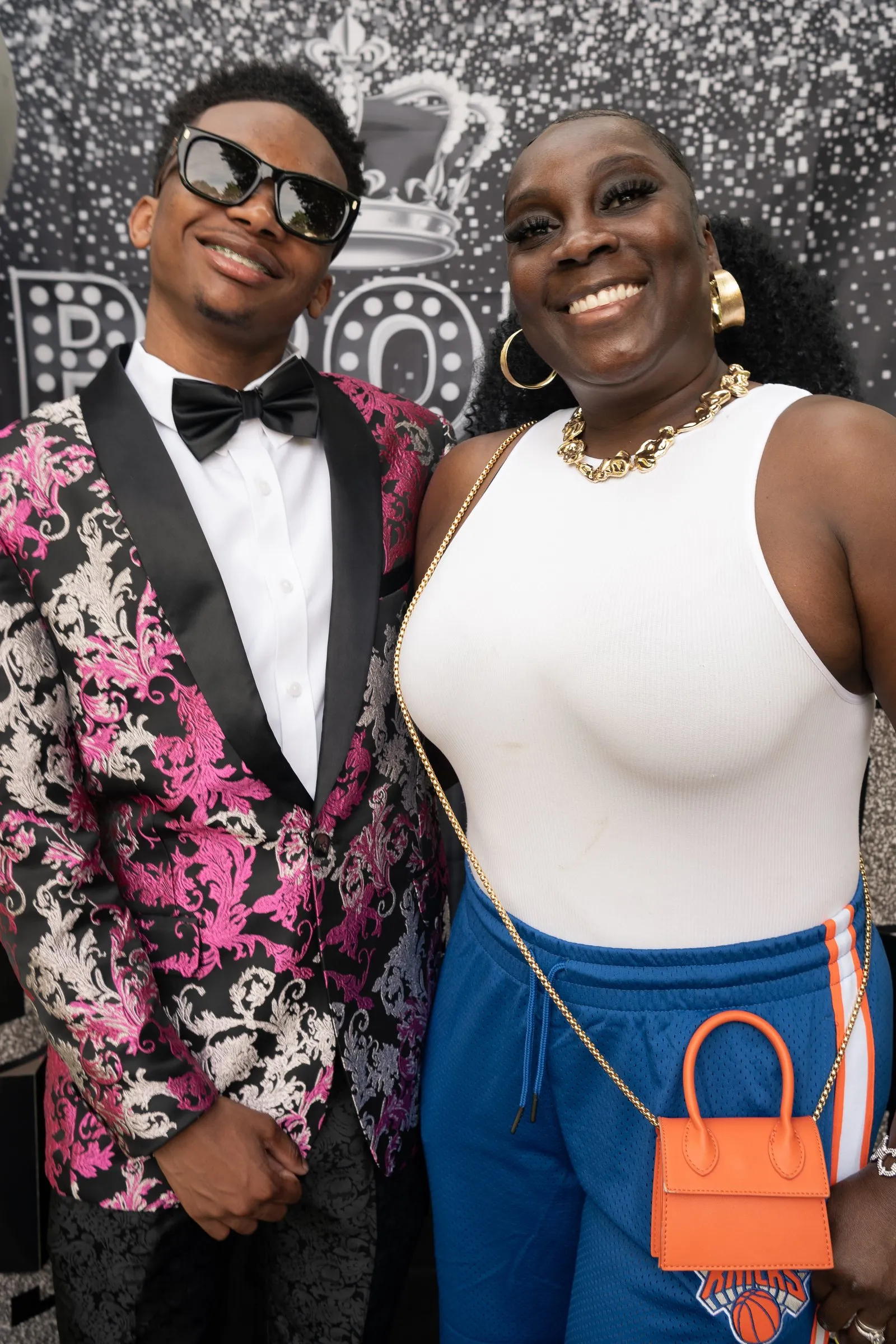 He poses with a woman in a white top and an orange handbag at the backdrop