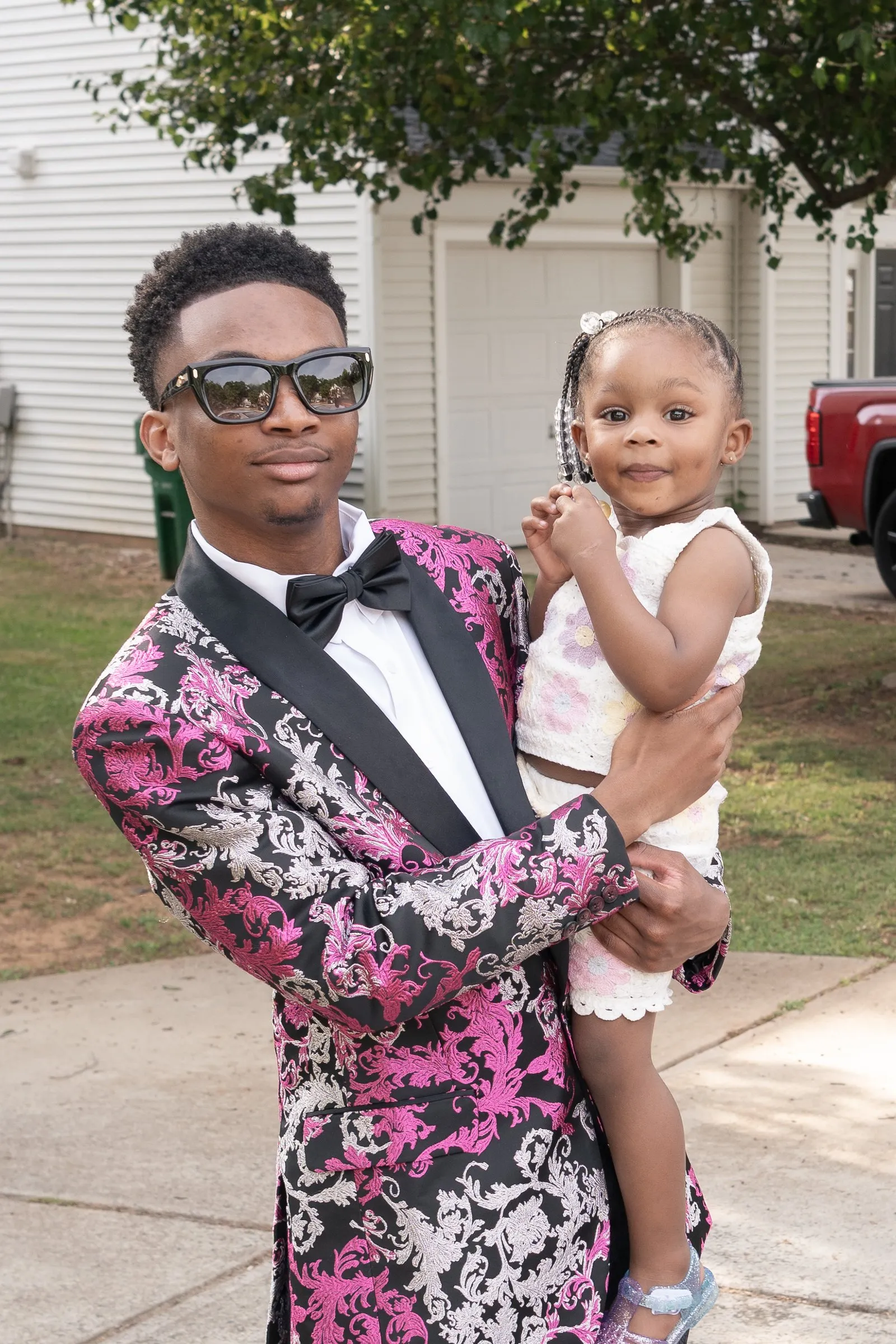 He holds a toddler on the front walk, both looking at the camera
