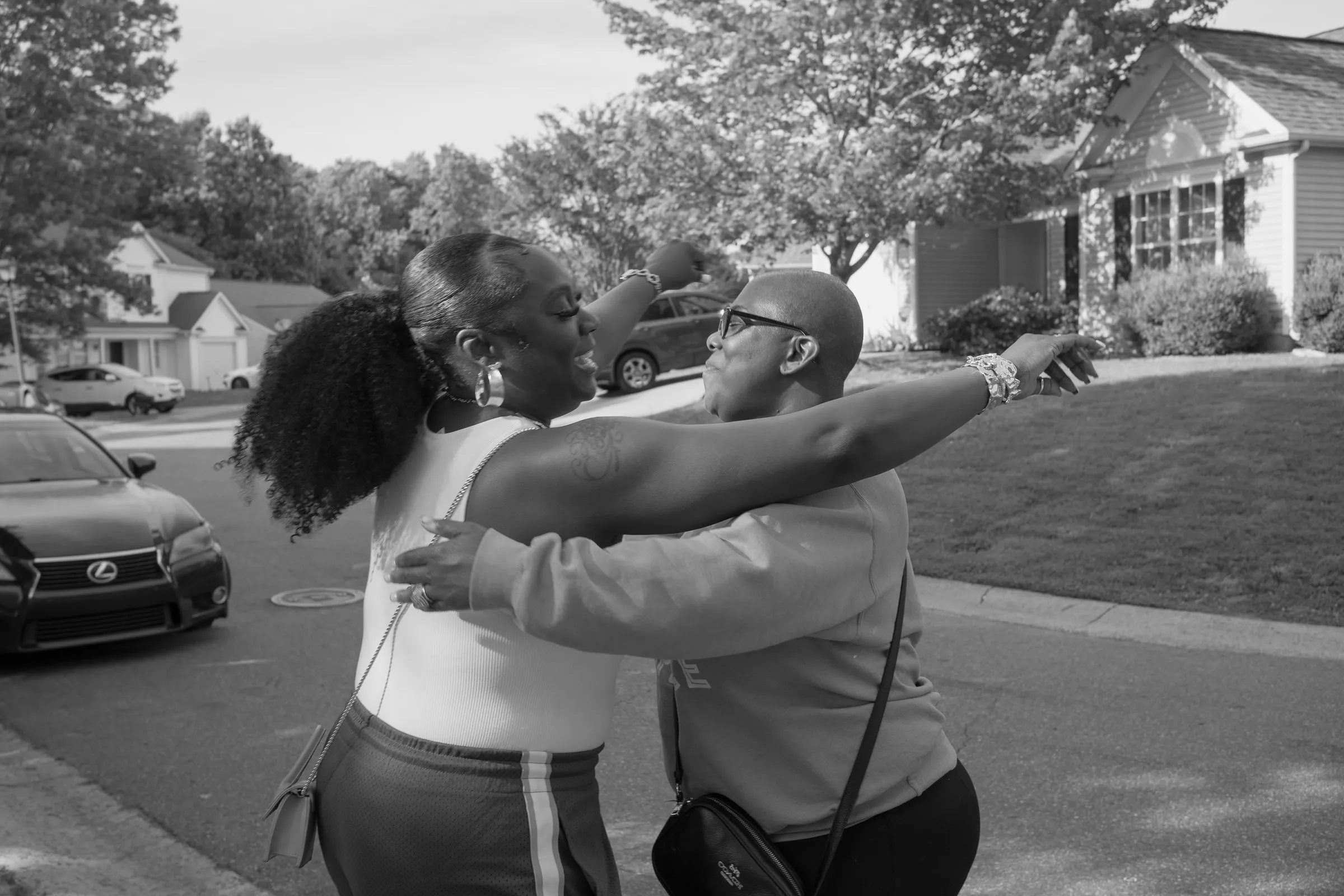 Black and white photograph of two guests hugging in the street as more family arrives