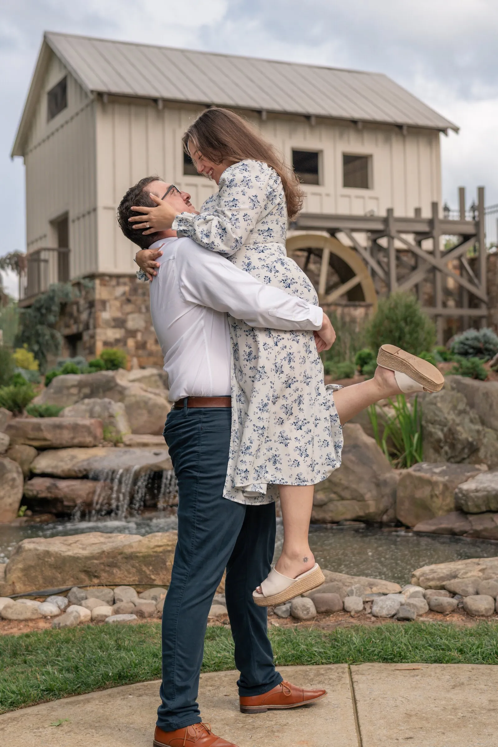 Spencer lifting Kayla off her feet beside the waterfall and white mill house at Millbridge in Waxhaw, NC
