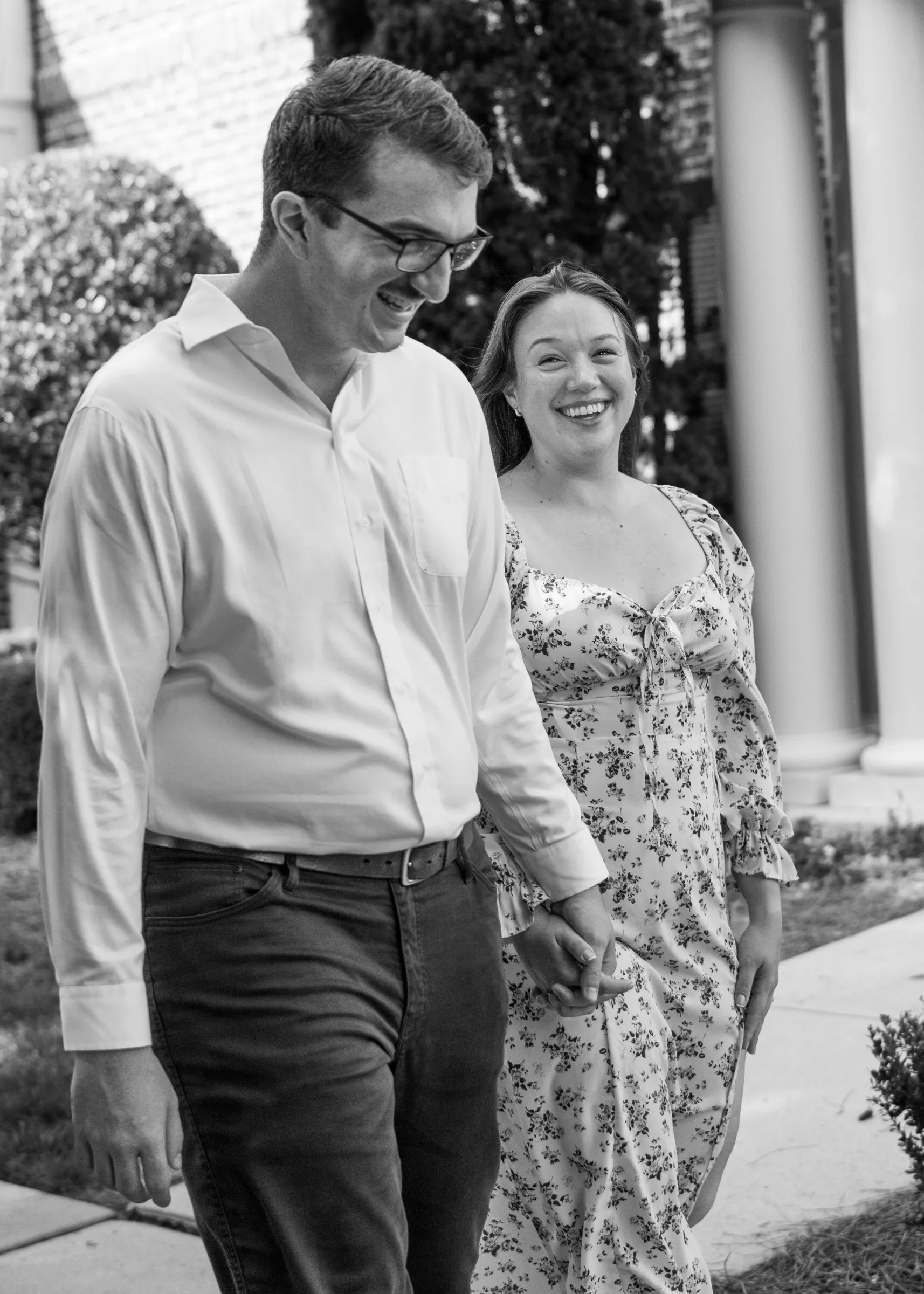 Black and white photograph of Spencer and Kayla laughing as they walk hand in hand past white columns