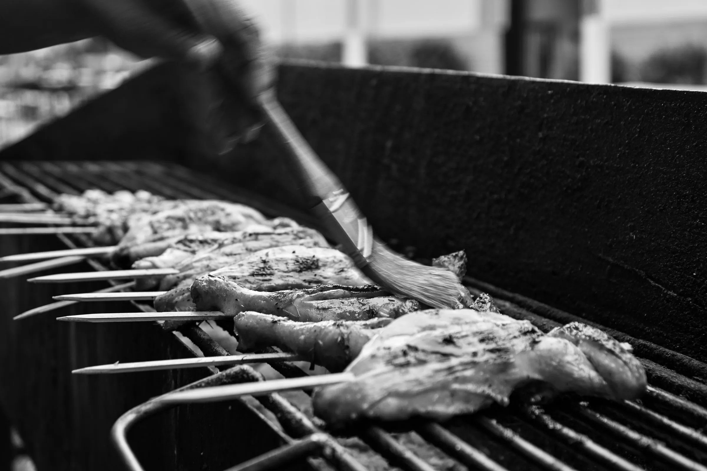 Black and white photograph of a basting brush glazing chicken skewers on the grill