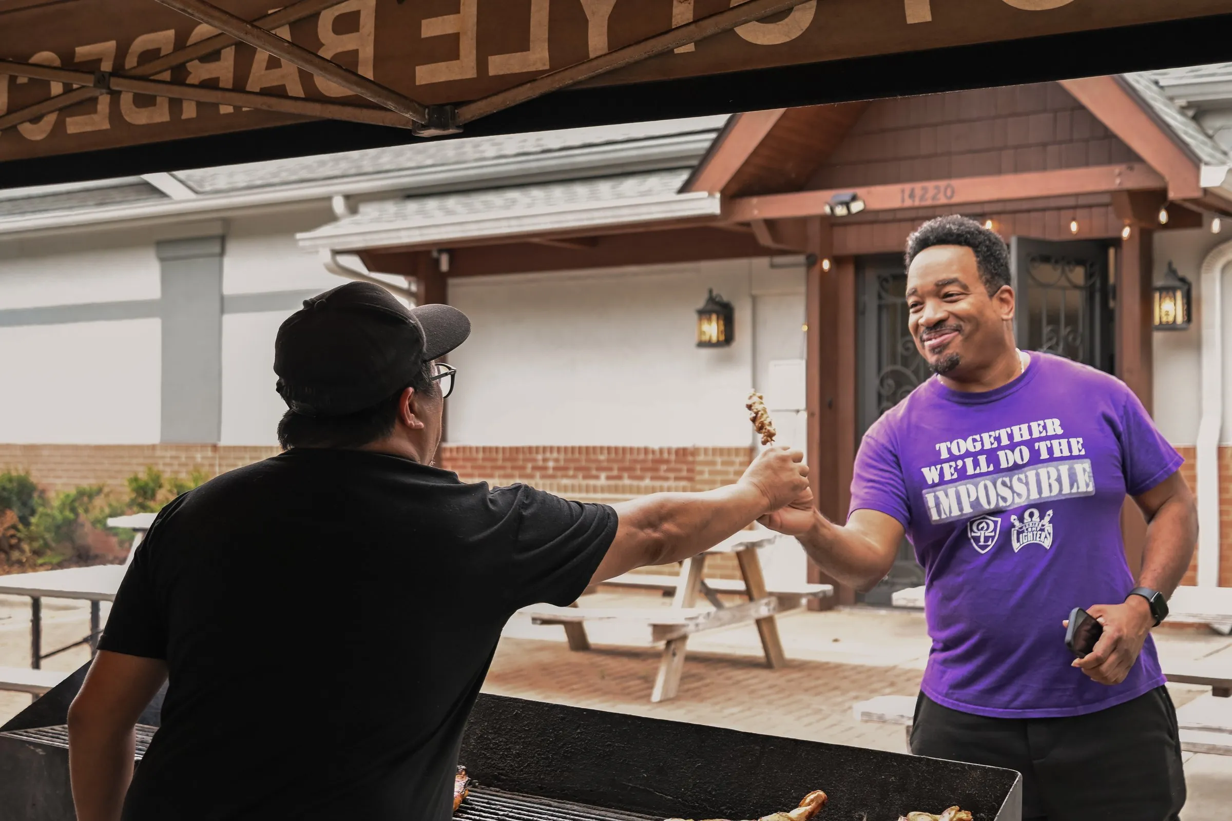 The cook hands a skewer across the grill to a smiling customer in a purple shirt