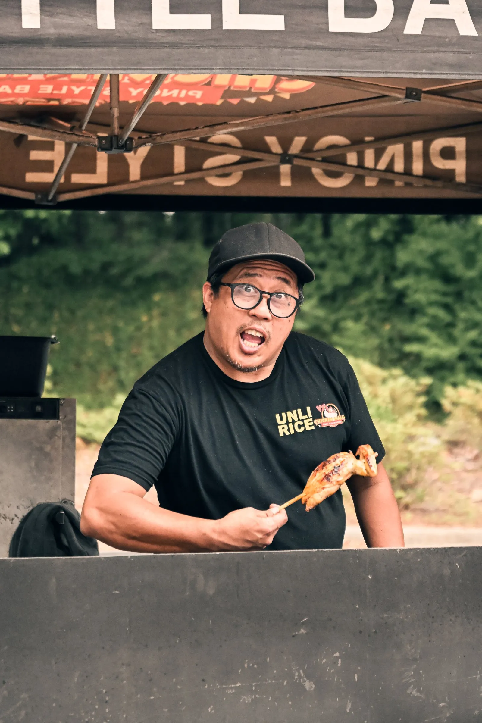 The cook in a black Unli Rice shirt shows off a skewer of grilled chicken with a surprised grin