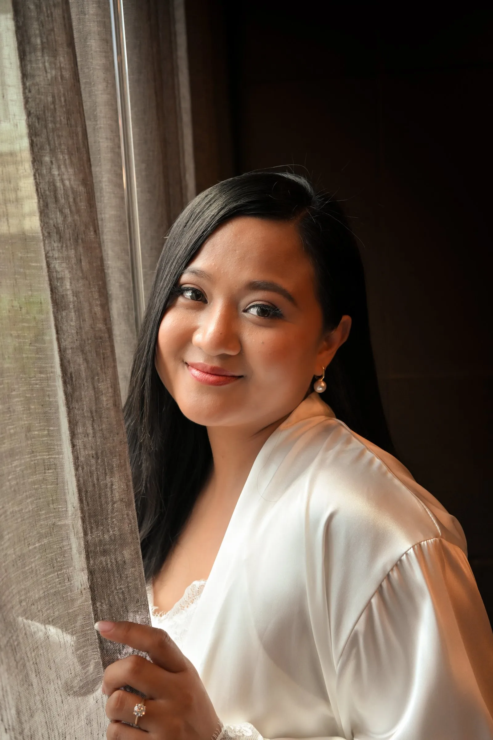 Window light bridal portrait of Winnie in her white satin robe, one hand on the curtain