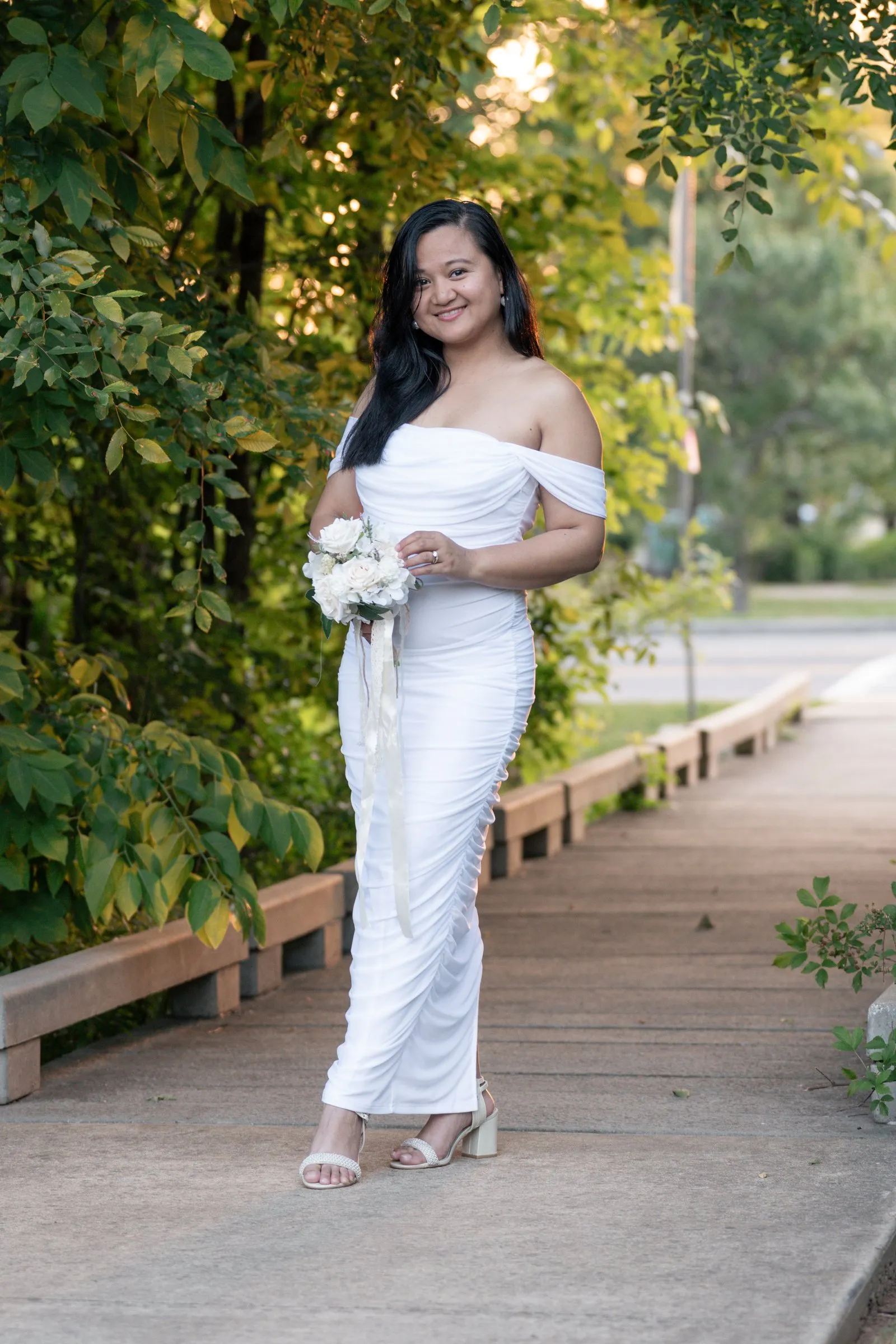 Winnie grinning on the wooden boardwalk in her white gown with green branches arching overhead