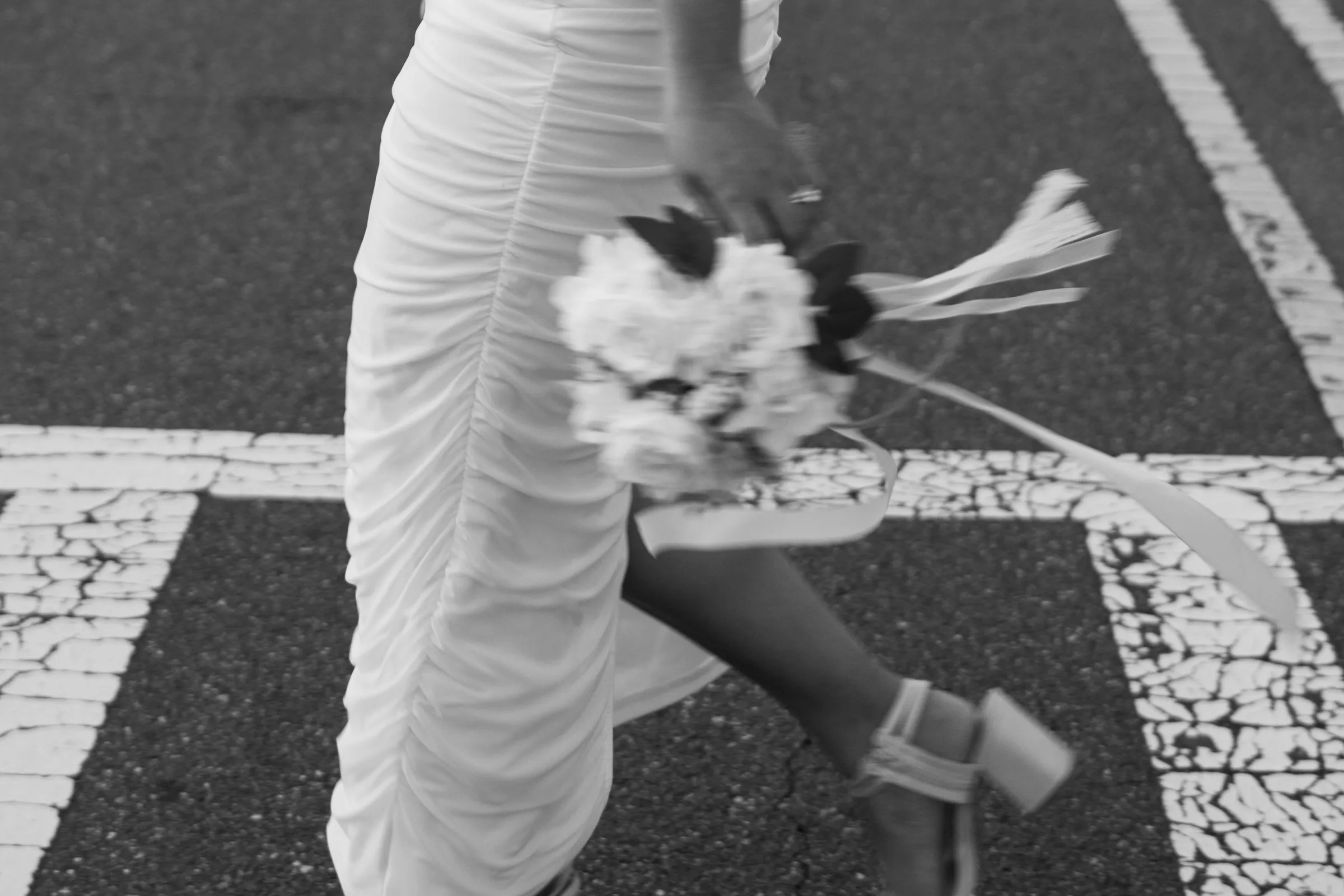 Black and white photograph of Winnie's gown and bouquet in motion as she steps across a weathered crosswalk