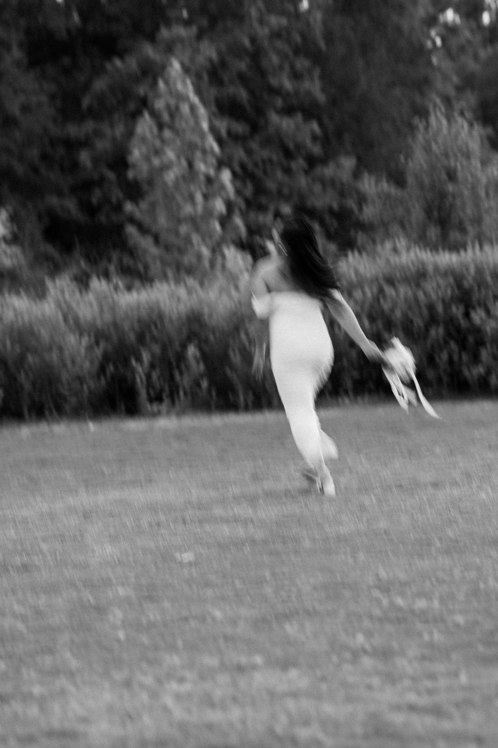 Black and white photograph of Winnie running across an open field toward the trees, her bouquet swinging in her hand