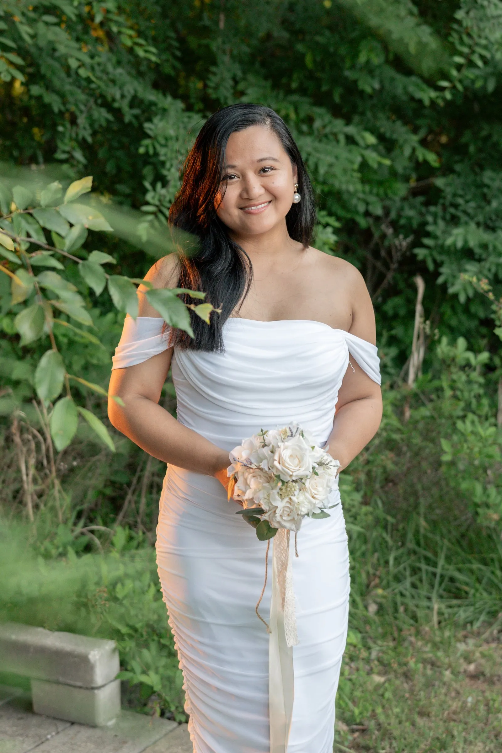 Winnie in her white gown among tall green plants, holding her rose bouquet with both hands