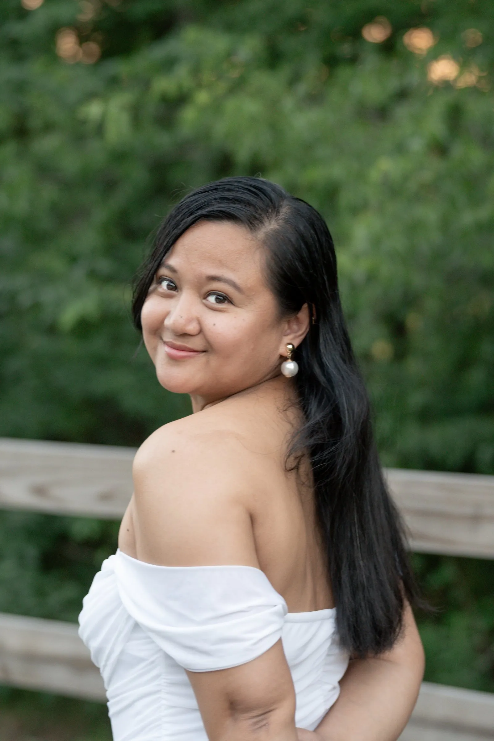 Winnie smiling back over her shoulder in front of the split rail fence, pearl earrings and dark hair over one shoulder