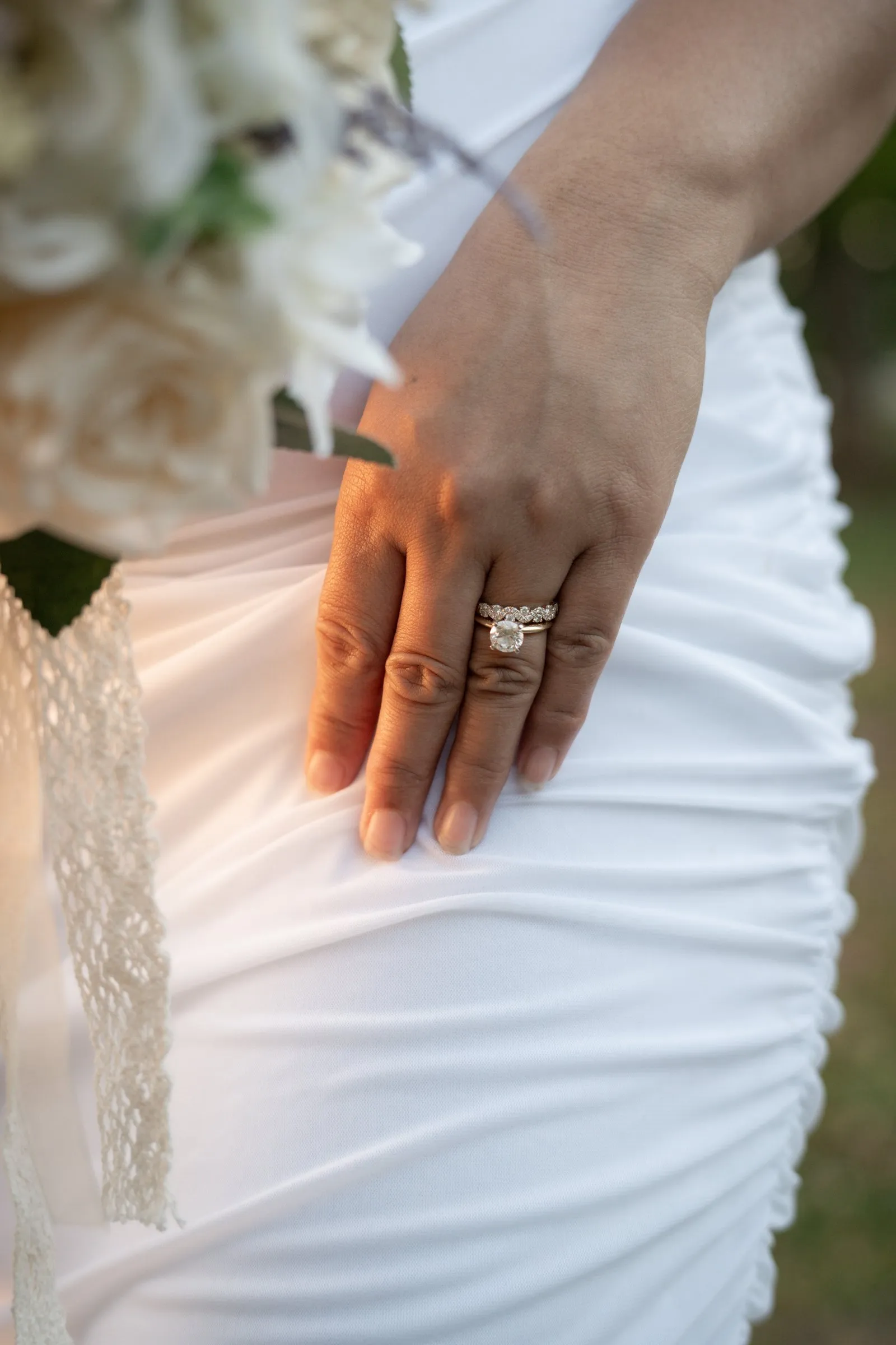 Detail of Winnie's hand resting on the ruched white gown, her engagement ring and wedding band catching the light