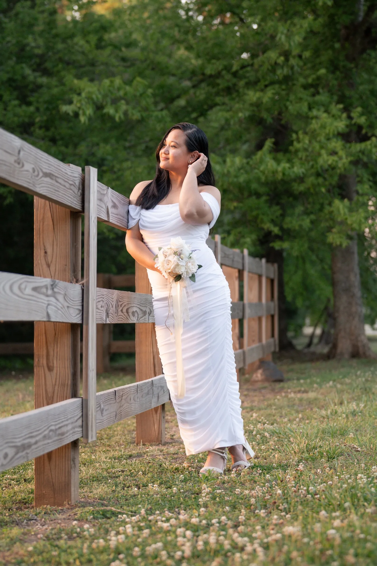 Winnie leaning against a wooden split rail fence in the grass, one hand in her hair, bouquet at her side