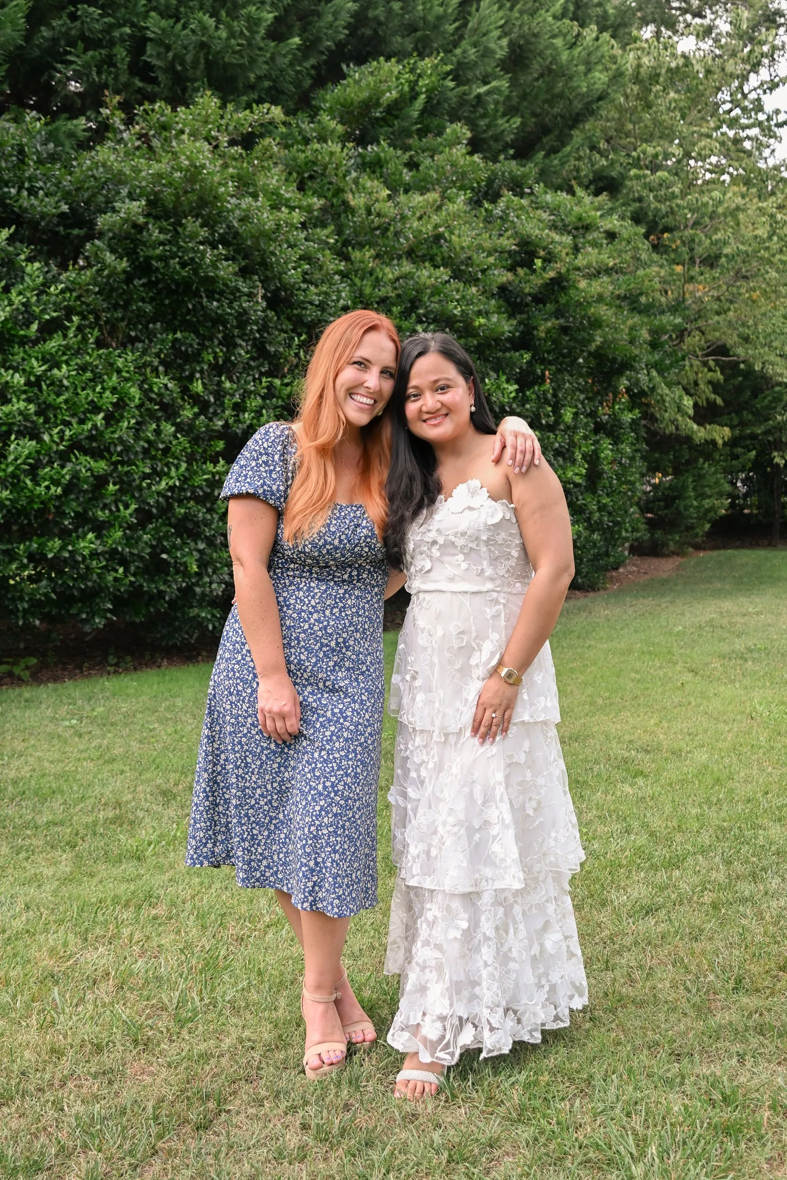 Winnie in a tiered white lace dress with a red haired bridesmaid in a blue floral dress, arm in arm on the lawn at Alexander Homestead