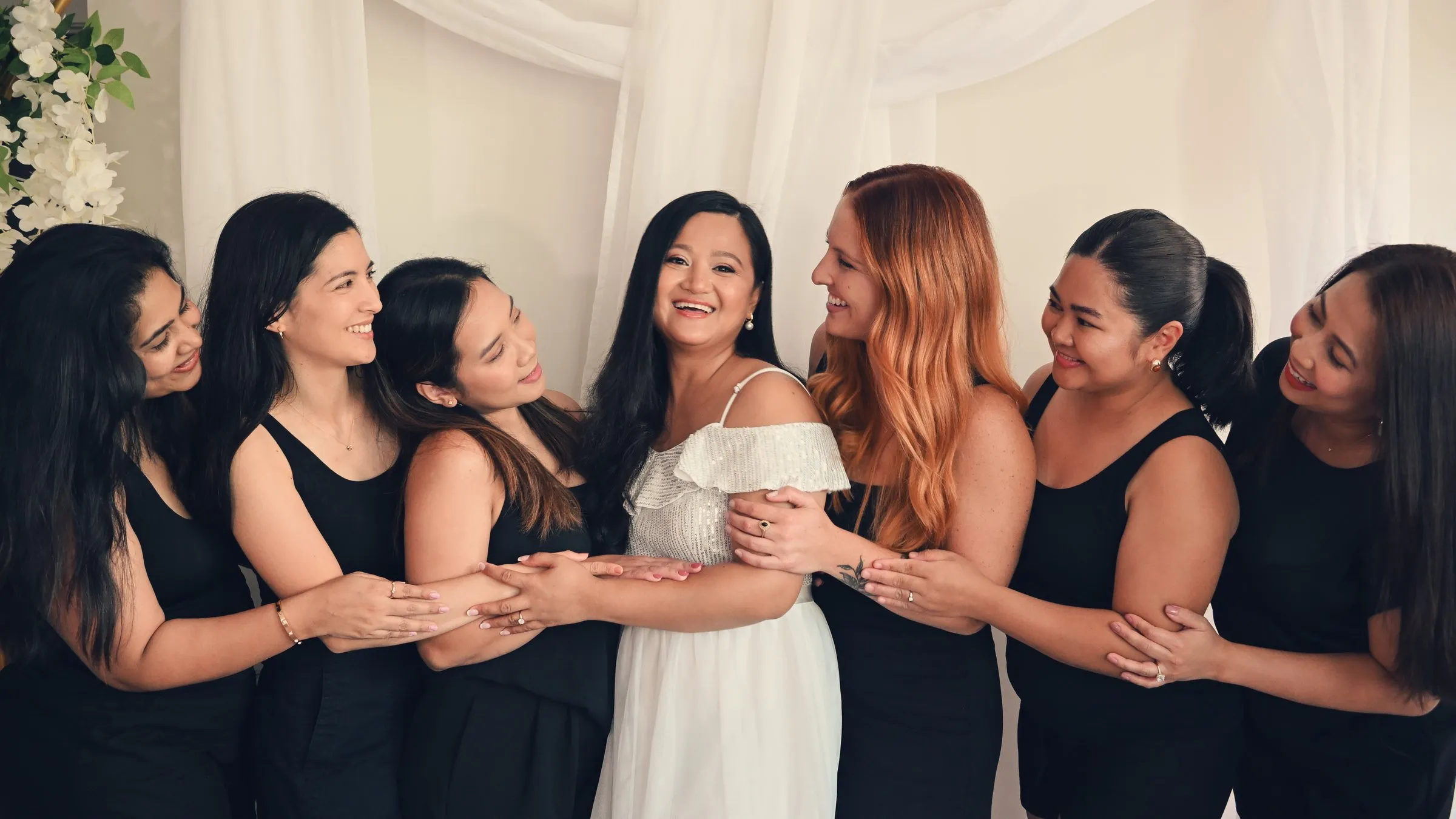 Winnie in white surrounded by six bridesmaids in black dresses, arms linked and everyone turned toward her, before the celebration dinner
