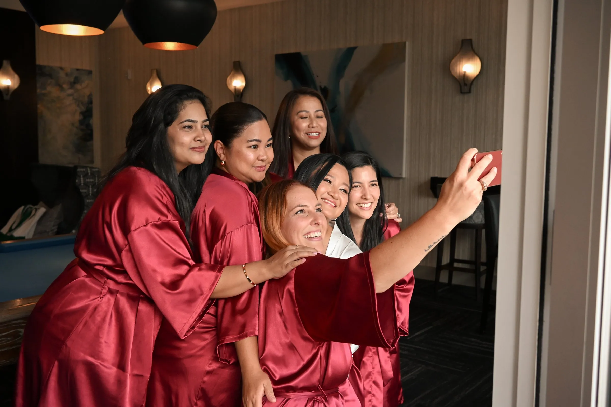 A bridesmaid taking a selfie with Winnie and the bridesmaids crowded in behind her, all in red satin robes