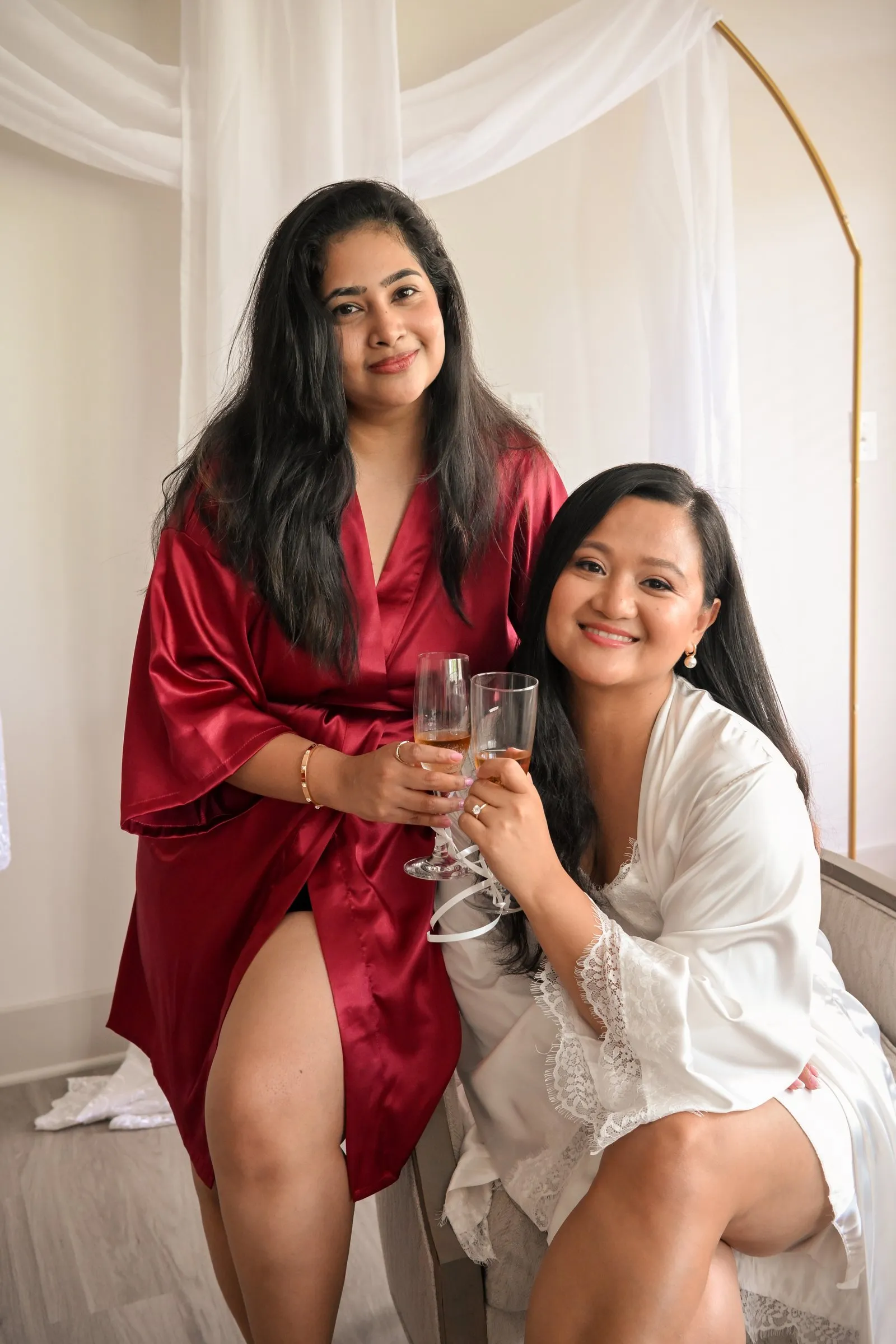 Winnie and her bridesmaid Sri seated together with champagne flutes, smiling at the camera